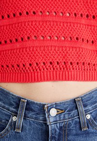 Rode gebreide crop top met opengewerkte patronen, gecombineerd met hoge blauwe denim jeans met een metalen knoop en contrasterende stiksels.