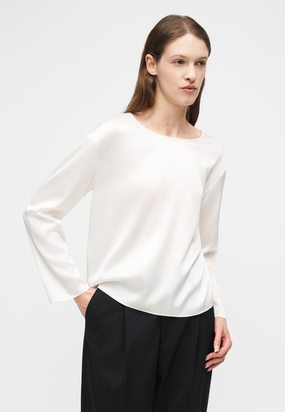 Γυναίκα που φοράει μια λευκή μακριά μπλούζα με άνετη εφαρμογή και μαύρο παντελόνι με φαρδύ πόδι, στέκεται με το ένα χέρι στην τσέπη μπροστά σε απλό φόντο.