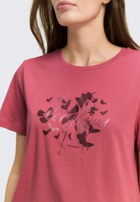 Roze katoenen T-shirt met een bloemmotief in donkere tinten en de tekst "Les Fleurs" in een metallic afwerking midden op het shirt.