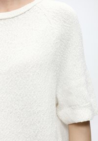 Κοντινό πλάνο ενός ατόμου που φοράει λευκό πλεκτό πουλόβερ με κοντά μανίκια, στρογγυλή λαιμόκοψη και υφασμάτινη υφή.