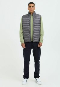 Chaleco acolchado gris con cuello alto, llevado sobre una camisa de manga larga verde claro, combinado con pantalones oscuros y zapatillas blancas con rayas negras.