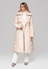 Žena v béžovém kabátu po kolena s kožešinovým límcem, bílém roláku, bílých kalhotách a kotníkových botách s hadím vzorem na jednobarevném pozadí.