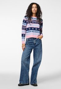 Pull en laine avec des rayures roses et bleu marine, featuring des motifs de texte et de flocons de neige. Pantalon en denim bleu large et bottes noires.