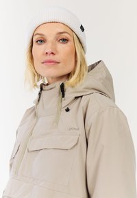 Hellebeige, wasserdichte Jacke mit hohem Kragen, Reißverschluss vorne, zwei Brusttaschen und verstellbarer Kapuze. Weiße, gerippte Strickmütze.