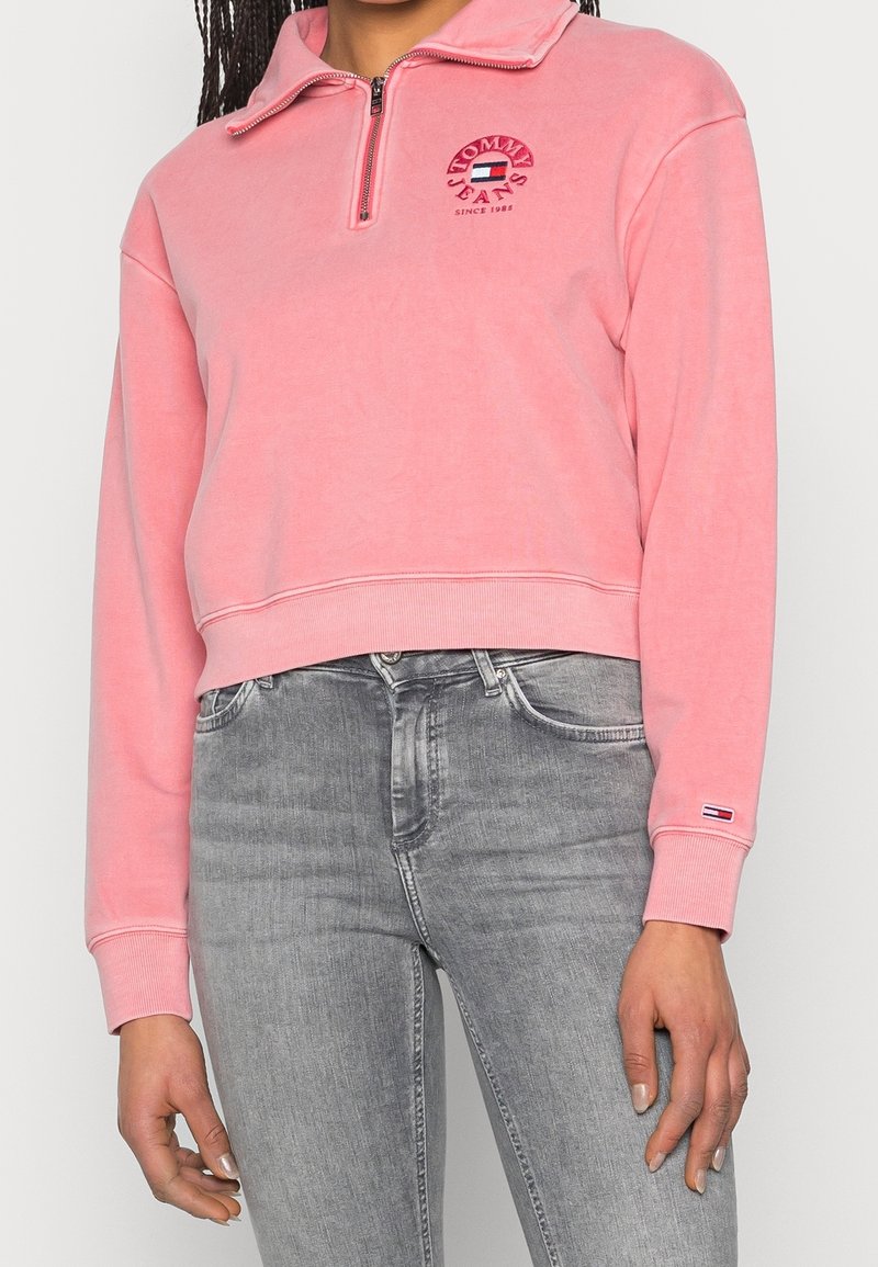 Pullover in pile rosa con collo a zip, presenta un logo rotondo sul petto. Polsini e orlo a coste, indossato sopra jeans grigi.