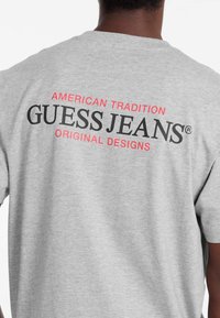 Pilkas medvilnės marškinėliai su juodu ir raudonu tekstu ant nugaros: "AMERIKIETIŠKA TRADICIJA," "GUESS JEANS®" ir "ORIGINALŪS DIZAINAI." Trumpi rankoviai.