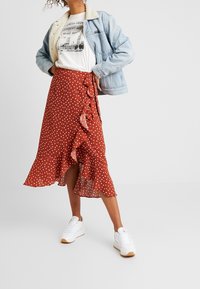 Gonna midi color ruggine a pois con orlo arricciato e fiocco laterale; abbinata a una t-shirt con stampa e una giacca di jeans, indossata con sneakers bianche.