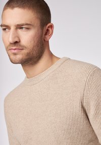 Beige gestrickter Pullover mit Rundhalsausschnitt, aus strukturiertem Stoff, mit dezenten Nähdetails. Nahaufnahme des Oberkörpers.