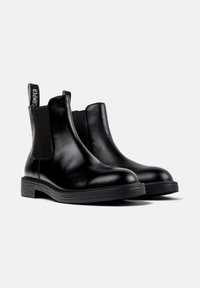 Une paire de bottes Chelsea en cuir noir avec des panneaux élastiques sur les côtés et des languettes de tirage avec logo, présentée sur un fond blanc uni.