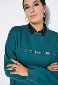 Felpa verde acqua con collo a giro, decorata con gemme colorate sul petto. Camicia nera con colletto sotto, orecchini a cerchio argentati.