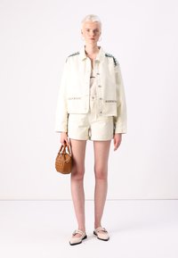 Chaqueta de mezclilla blanca con acentos verdes, combinada con pantalones cortos blancos. Sostiene un pequeño bolso tejido marrón y lleva mocasines beige.