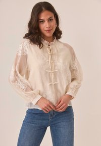 Mujer con cabello oscuro y ondulado vestida con una blusa sheer color crema con bordados florales y jeans azules, de pie frente a un fondo beige.