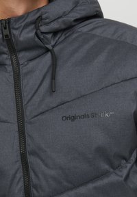 Grå pufferjakke med en tekstureret stof, med lynlås foran, justerbar hætte og "Originals Studio" branding på brystet.