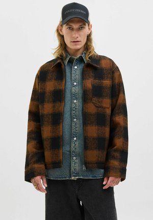 Young man wearing a black and brown checkered jacket over a blue denim shirt and a black Pasadena cap, standing against a plain background.