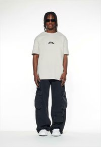 Šviesiai pilkas per didelis marškinėliai su juodu ir mėlynu užsiuviniu "YOUTH", derinami su juodomis kargo kelnėmis, turinčiomis kelis kišenes, ir baltais sportiniais bateliais.