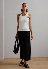 Witte geribbelde mouwloze top met zwarte accenten, gecombineerd met zwarte wijde broek en zwarte instapsandalen. Het model houdt een zwarte totebag vast.