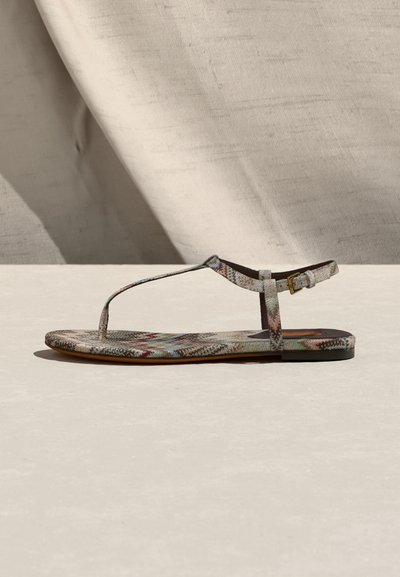 Επίπεδη σάνδαλα με πολύχρωμο γεωμετρικό μοτίβο, λεπτό Τ-λουράκι και πλαϊνή αγκράφα, παρουσιάζεται πάνω σε ουδέτερο υπόστρωμα με απαλό φόντο ύφασμα.