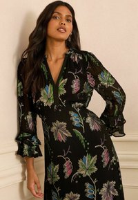 Schwarzes Kleid mit bunten Blumenstickereien in Grün, Pink und Blau. Es hat einen V-Ausschnitt und durchsichtige Rüscheärmel.