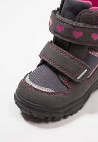 Šedá dětská bota s růžovými vzory srdcí a suchými zipy, vybavená protiskluzovou podrážkou a texturovaným materiálem na bílém pozadí.