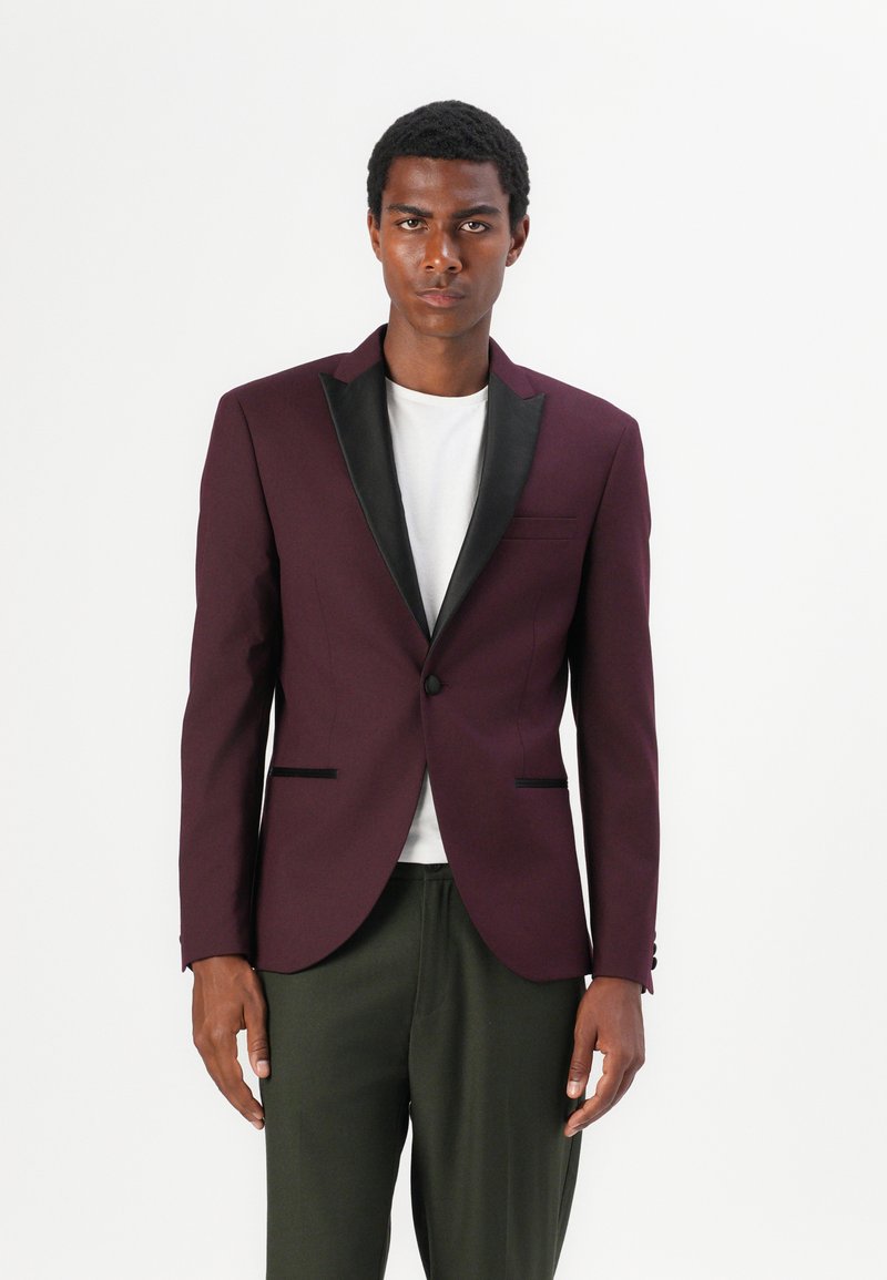Bordeauxfarbener Blazer mit schwarzen Satinrevers und Taschenbesatz, einreihiger Knopfverschluss, taillierter Schnitt, getragen über einem weißen Hemd und grünen Hosen.