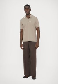 Beige polo shirt with striped collar, paired with dark brown pleated trousers. Model stands against a light background, showcasing outfit details.