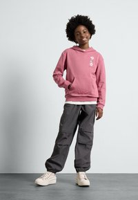 Sweat à capuche rose avec poche avant et motif graphique, associé à un pantalon cargo gris ample et des baskets beige.