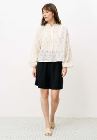 Spitzenbluse in Off-White mit floralem Muster, mit einem knopfgeknöpften Ausschnitt und puffy Ärmeln, kombiniert mit einem schwarzen Rock und hautfarbenen Sandalen.