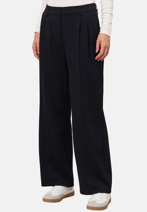 Pantalons larges bleu marine avec un devant plissé, fabriqués dans un tissu doux. Présentent des poches avant et une taille ajustée, associés à des baskets blanches.