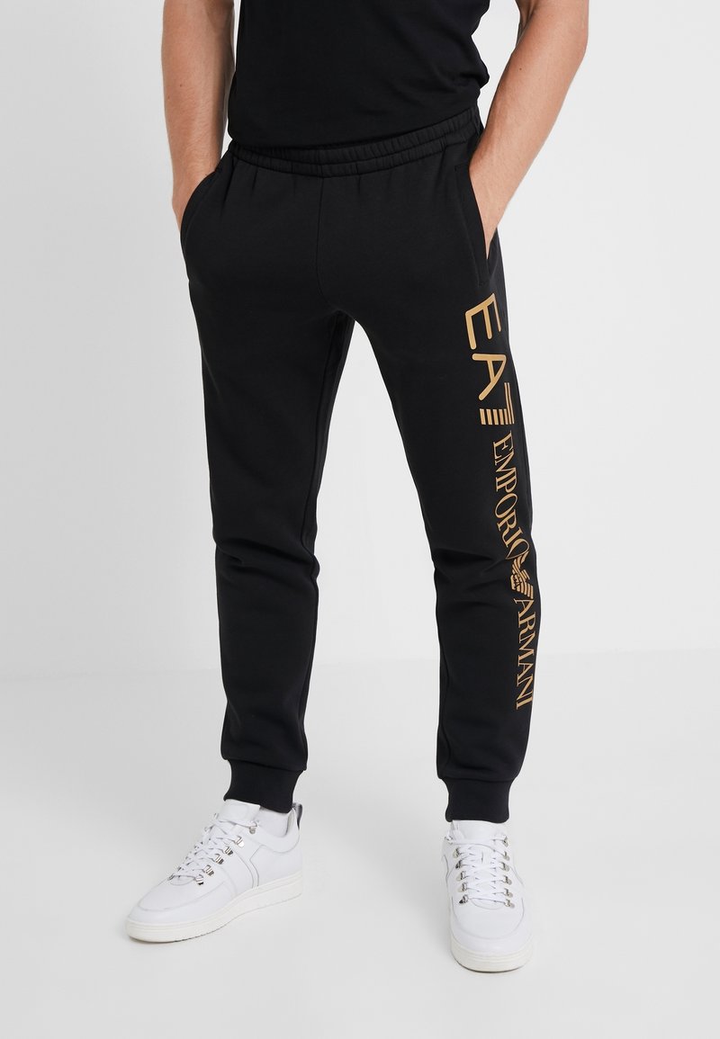 Muž v černých joggingových kalhotách s zlatým logem "EA7 Emporio Armani" na levé nohavici, bílých teniskách a černém tričku, se rukama v kapsách.