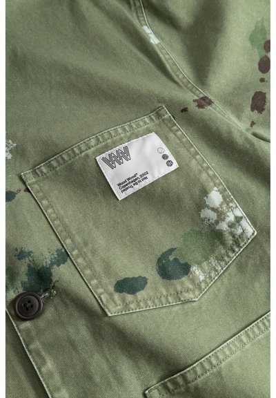 Camisa de algodón en verde oliva con un bolsillo de parche, que presenta una etiqueta y salpicaduras de pintura verde oscuro y marrón. Tejido texturizado.