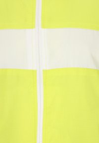 Kirkas keltainen ja valkoinen korkeavisiivinen takki etuvetoketjulla. Kankaan textuuri sisältää vaakasuoria väripaloja näkyvyyden parantamiseksi.