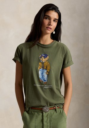 Femme portant un T-shirt vert olive avec un motif d'ourson en lunettes de soleil, veste et jean, rentrée dans un pantalon taille haute avec une boucle de ceinture décorative.