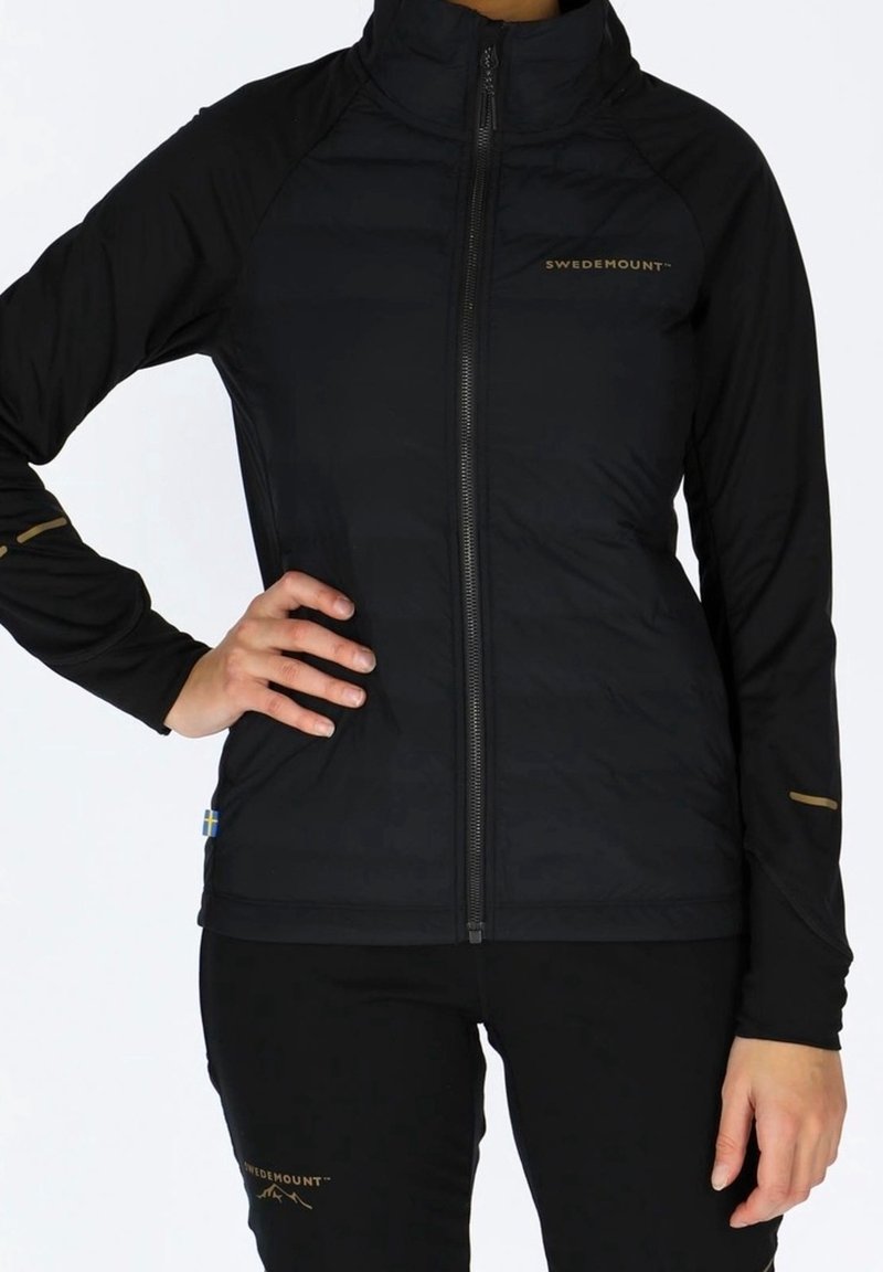 Veste noire zippée avec une texture matelassée et une coupe ajustée. Logo doré sur la poitrine et les manches. Associée à un pantalon noir.