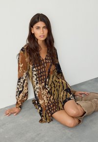 Abito multicolore in marrone, nero e beige con motivi astratti, maniche lunghe e scollo a V, abbinato a stivali beige al ginocchio.