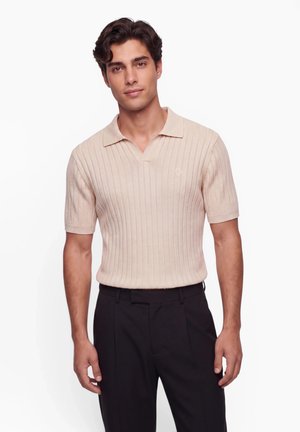 Man wearing a beige short-sleeve ribbed knit polo shirt and black trousers standing against a white background.