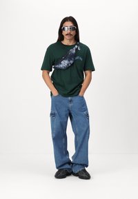 Grøn bomulds t-shirt med lomme, blå cargo jeans, sort crossbody taske og reflekterende solbriller. Løst og afslappet urban outfit.