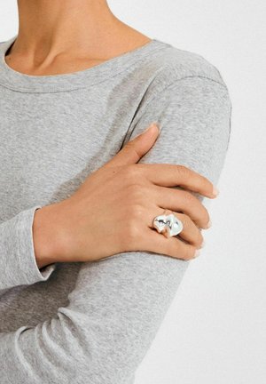 Hõbedast metallist sõrmus skulpturaalse lainelise disainiga, mis on kantud käel. Käsi toetub hallile pikkade varrukatega särgile, tuues esile sõrmuse tekstuuri.