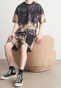 Persona sentada en una silla beige texturizada, vistiendo pantalones cortos de teñido anudado en negro y marrón y una camiseta, con zapatillas altas negras y calcetines negros.