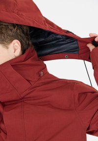 Rote Außenjacke mit hohem Kragen, verstellbarem Kapuze und dunkler Innenauskleidung. Verfügt über Reißverschluss und Druckknopfverschlüsse sowie strukturierter Stoff.