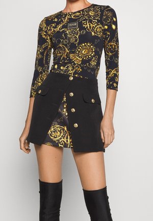 Haut à manches longues noir et or à motif avec logo visible, associé à une mini-jupe noire ornée de boutons dorés et des cuissardes noires.