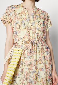 Robe sans manches jaune avec un imprimé de papillons multicolores, taille à cordon, boutons, et une personne tenant un sac tissé rayé jaune et blanc.