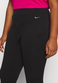 Sorte træningsleggings med en bred talje, som har en sidelomme og et lille reflekterende logo på venstre hofte. Glat, strækbart stof.