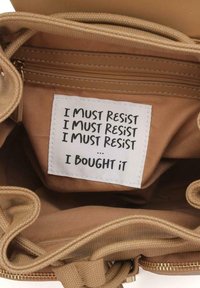 Intérieur de sac en tissu beige avec une étiquette portant un texte noir : "JE DOIS RÉSISTER / JE DOIS RÉSISTER / JE DOIS RÉSISTER / ... JE L'AI ACHETÉ. "