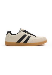 Zapatilla beige clara con acentos negros y tres rayas negras. Material suave con una puntera redondeada y una suela de goma color caramelo. Cintas negras.