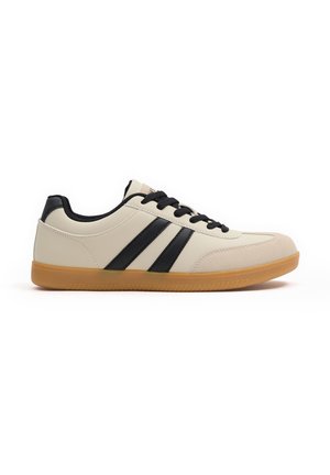 Zapatilla beige clara con acentos negros y tres rayas negras. Material suave con una puntera redondeada y una suela de goma color caramelo. Cintas negras.