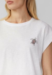 Biała bawełniana koszulka z kolorowym haftem żółwia na lewej piersi. Projekt zawiera skomplikowane wzory i miękką teksturę materiału.