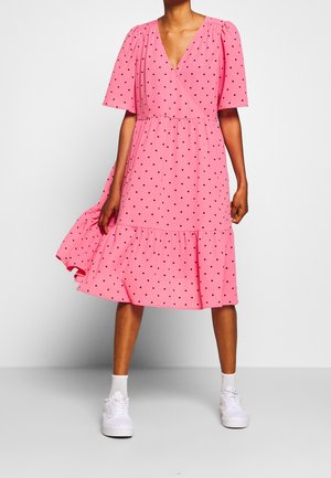 Kvinde iført en lyserød knælængde kjole med sorte polkaprikker, korte ærmer, V-hals og hvide sneakers med sokker, stående foran en ensfarvet baggrund.