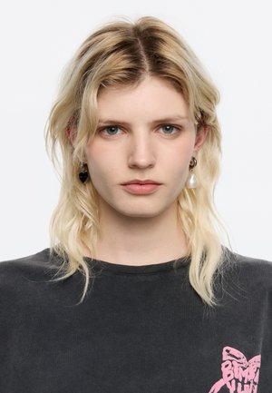Mood with lainete blonde hair, wearing a gray shirt with a pink graphic. Accessories include black heart earrings and a pearl drop earring.