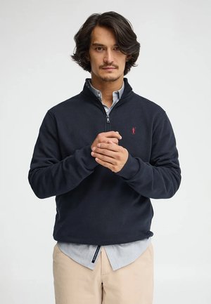 Uomo con capelli scuri di media lunghezza che indossa una felpa blu navy con mezza zip sopra una camicia azzurro chiaro e pantaloni beige, in piedi davanti a uno sfondo bianco neutro.