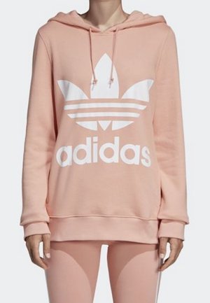 Person trägt einen hellrosa Adidas-Hoodie mit großem weißem Trefoil-Logo und passende Leggings.